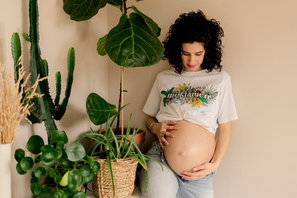 Femme enceinte portant un jean ouvert et un t-shirt remonté sous la poitrine à côté de plusieurs plantes d'intérieur