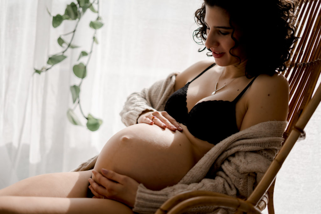 Femme enceinte assise dans un fauteuil en rotin et portant un gilet beige ouverte sur de la lingerie noire pendant sa séance photo maternité à Niort