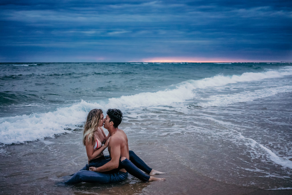 Un couple s'embrasse assis au bord des vagues devant le coucher du soleil pendant leur séance photo dans les Landes
