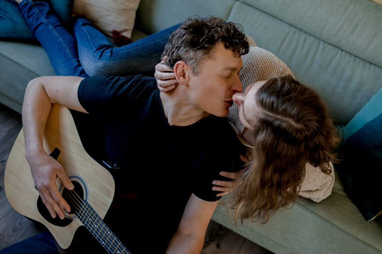 Séance photo de couple avec un homme et une femme s'embrassant. L'homme joue de la guitare. La femme est allongée sur un canapé.